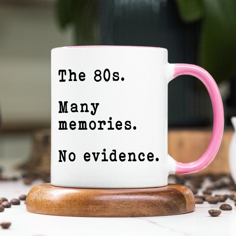 May include: White coffee mug with a pink rim and handle. The mug has black text that reads "The 80s. Many memories. No evidence."