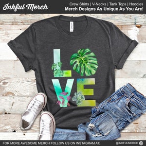 May include: A dark gray t-shirt with a watercolor design of the word "LOVE" made up of green leaves. The shirt is laid flat on a wooden surface with a pair of white sneakers and blue jeans.