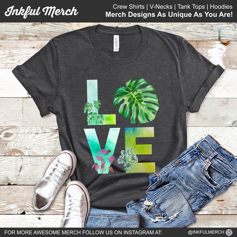 May include: A dark gray t-shirt with a watercolor design of the word "LOVE" made up of green leaves. The shirt is laid flat on a wooden surface with a pair of white sneakers and blue jeans.