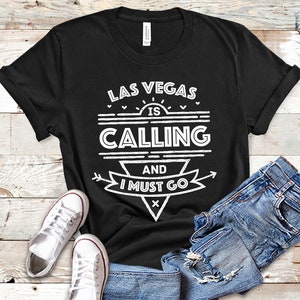 Pu&ograve; includere: Maglietta nera con testo bianco che recita "Las Vegas is Calling and I Must Go". Il testo &egrave; in un font in stile vintage con un sole e frecce.