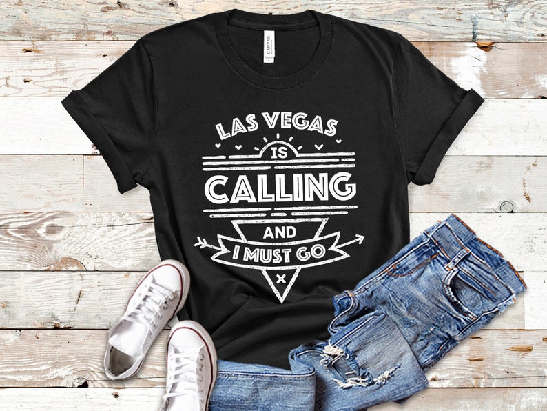 Pu&ograve; includere: Maglietta nera con testo bianco che recita "Las Vegas is Calling and I Must Go". Il testo &egrave; in un font in stile vintage con un sole e frecce.