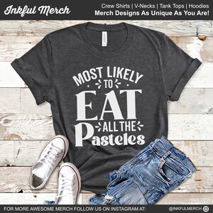 May include: A dark gray t-shirt with white text that reads "Most Likely To Eat All The Pasteles". The shirt is displayed on a wooden surface with a pair of white sneakers and blue jeans.