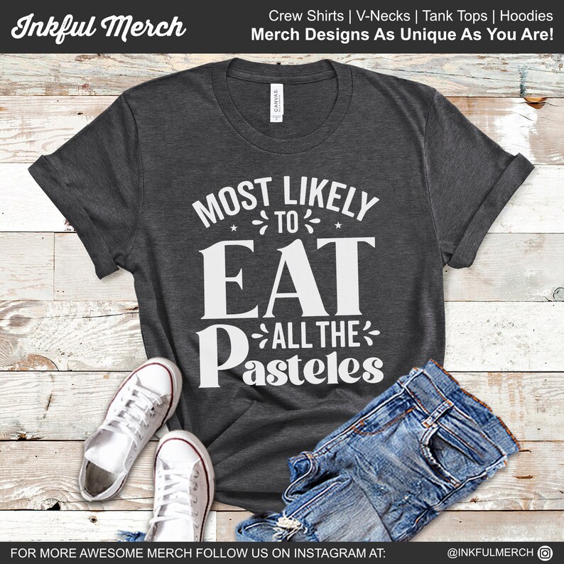 May include: A dark gray t-shirt with white text that reads "Most Likely To Eat All The Pasteles". The shirt is displayed on a wooden surface with a pair of white sneakers and blue jeans.