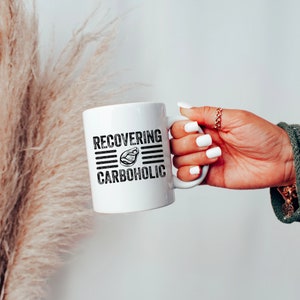 May include: White ceramic mug with black text that reads "RECOVERING CARBOHOLIC" with a graphic of a steak in the middle of the text.