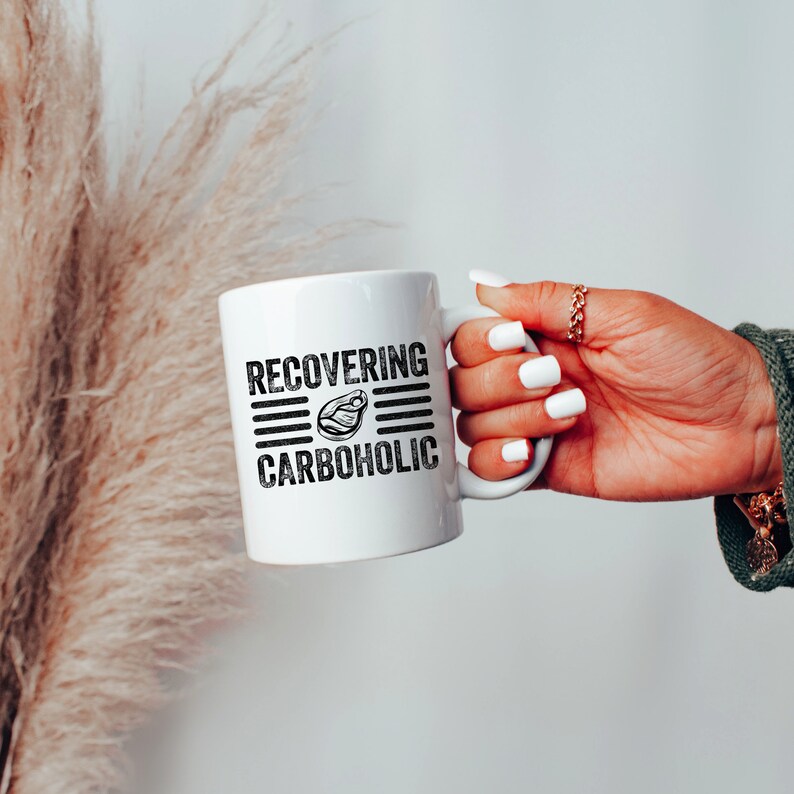 May include: White ceramic mug with black text that reads "RECOVERING CARBOHOLIC" with a graphic of a steak in the middle of the text.
