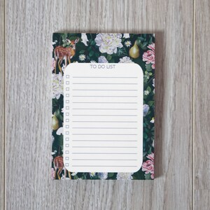 Peut inclure: Un bloc-notes blanc à faire avec un motif floral et de cerfs. Le bloc-notes a un fond vert avec des fleurs blanches, roses et jaunes, une poire et un cerf.