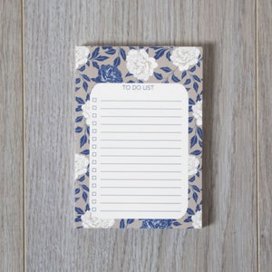 Könnte beinhalten: Ein weißer Notizblock für To-Do-Listen mit einem floralen Design mit blauen Blättern und weißen Blüten. Der Notizblock hat einen blauen Rand und die Worte "TO DO LIST" oben aufgedruckt.