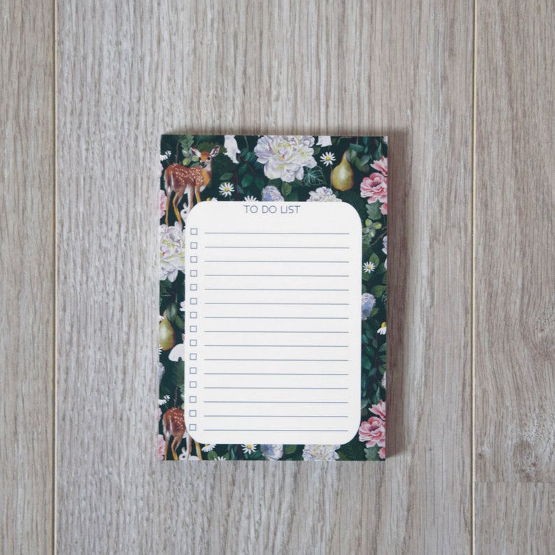 Pu&ograve; includere: Un blocco note bianco da fare con un design floreale e di cervi. Il blocco note ha uno sfondo verde con fiori bianchi, rosa e gialli, una pera e un cervo.