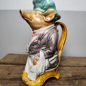 May include: A ceramic pitcher shaped like a pig wearing a chef's hat and uniform. The pig is tan with a blue hat, a pink bow tie, and a gray jacket. The pitcher is seated on a yellow chair with a handle.