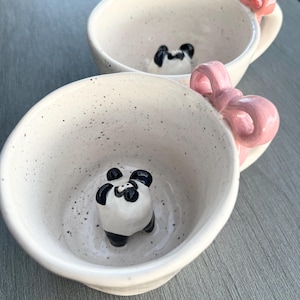 Könnte beinhalten: Zwei weiße Keramikbecher mit rosa Schleifen und einer kleinen Pandabärenfigur in einem der Becher.