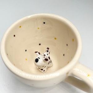 Op de afbeelding: Een witte keramische mok met een bruin en geel gespikkeld ontwerp. In de mok zit een klein keramiek beeldje van een dalmatiër.