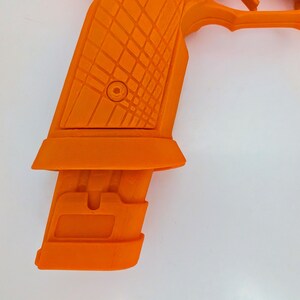 May include: Bright orange handgun with a textured grip and magazine. The grip features a diamond pattern, and the magazine is inserted into the handle. The gun is set against a white background.