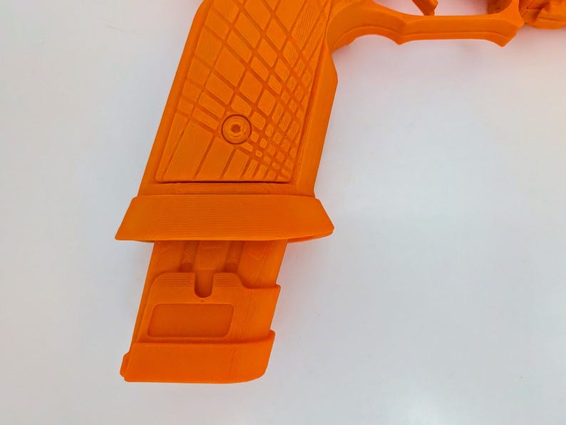 May include: Bright orange handgun with a textured grip and magazine. The grip features a diamond pattern, and the magazine is inserted into the handle. The gun is set against a white background.