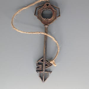 May include: A vintage-style key with a decorative design, suspended by a natural twine string. The key has a bronze finish and features an ornate head and a stylized lower portion. The key is approximately 6 inches long.