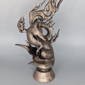 May include: A bronze fox sculpture with flame-like details. The fox is depicted in a seated pose, with its tail and the flames extending upwards. The sculpture has a textured surface and is mounted on a pedestal base.