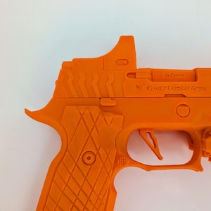 May include: Bright orange handgun with a textured grip and a sight on top. The gun features a trigger, a slide, and the text "Kandy Tactical Arms". The overall design is angular and modern.