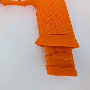 May include: Close-up of an orange handgun with a textured grip and magazine. The gun is set against a white background, highlighting its bright color and geometric design. The magazine is inserted into the handle.