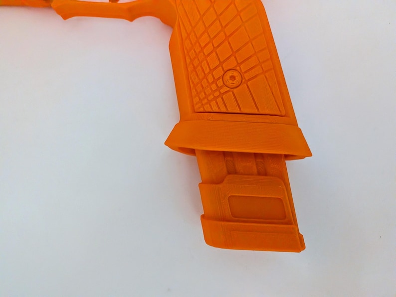 May include: Close-up of an orange handgun with a textured grip and magazine. The gun is set against a white background, highlighting its bright color and geometric design. The magazine is inserted into the handle.