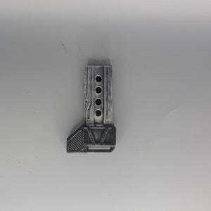 May include: A small, rectangular, metallic-looking object with a series of four circular holes. The object has a textured surface and a geometric design on one end. The color appears to be a muted gray or silver.
