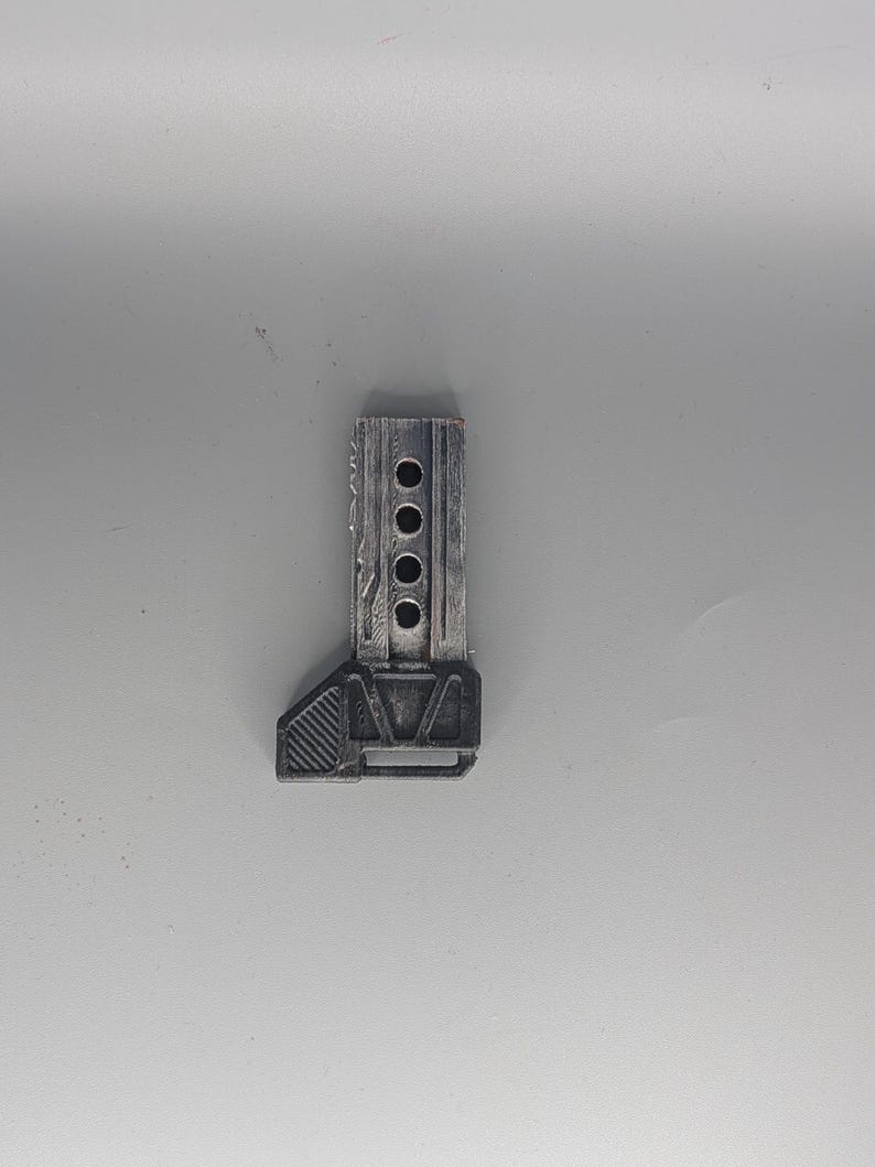 May include: A small, rectangular, metallic-looking object with a series of four circular holes. The object has a textured surface and a geometric design on one end. The color appears to be a muted gray or silver.