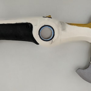 May include: A white and black prop weapon with a gold handle and a gray blade. The handle is black, and the blade is gray. The weapon has a circular opening and a small gold detail near the handle.