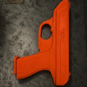 May include: A bright orange toy pistol featuring the word Matilda embossed on the side. The object has a textured surface and a distinct trigger guard. It is positioned against a dark, weathered metal surface with a rectangular label reading ITEM BOX.