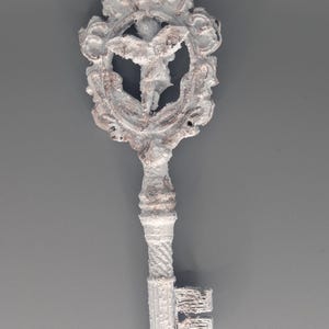 May include: Ornate, antique-style key with a white and gray finish. The key features a decorative head with a central figure within a detailed frame. The handle and teeth of the key are also intricately designed, suggesting a vintage or fantasy theme.