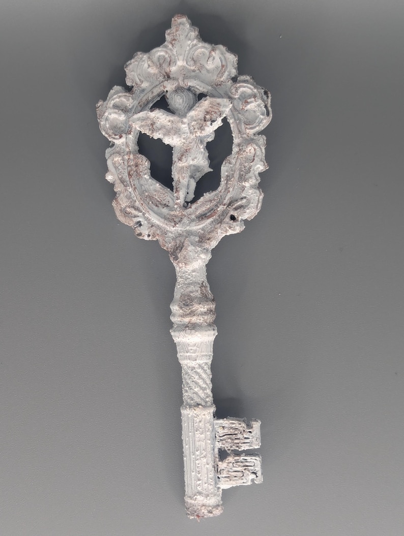 May include: Ornate, antique-style key with a white and gray finish. The key features a decorative head with a central figure within a detailed frame. The handle and teeth of the key are also intricately designed, suggesting a vintage or fantasy theme.
