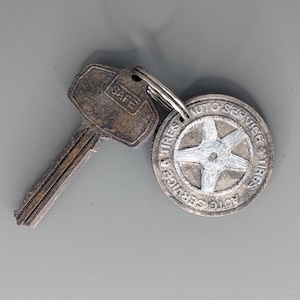 Op de afbeelding: Een vintage sleutel en een ronde metalen sleutelhanger. De sleutel is gestempeld met "SAFE". De sleutelhanger heeft de woorden "AUTO SERVICE TIRES VIRGS" rond een stervormig ontwerp. Beide zijn bevestigd aan een zilveren sleutelring.