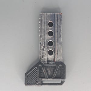 May include: A weathered, rectangular, gray and black object with four circular holes and a textured surface. The object has a geometric base and appears to be made of wood or a similar material. The design suggests a mechanical or industrial theme.