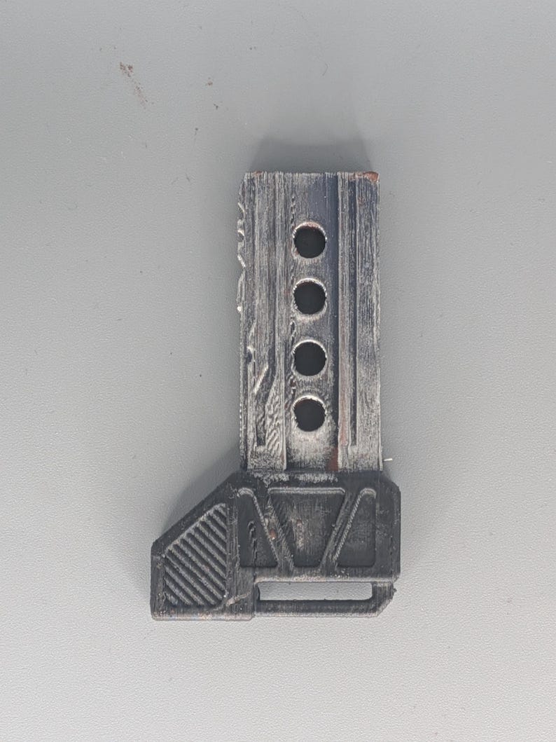 May include: A weathered, rectangular, gray and black object with four circular holes and a textured surface. The object has a geometric base and appears to be made of wood or a similar material. The design suggests a mechanical or industrial theme.