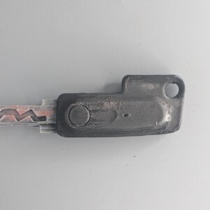 May include: A worn, dark gray key with a distinctive jagged edge and a rectangular head. The key has a circular indentation and a hole at the end, suggesting it's for a lock. The key's metal shows signs of wear.