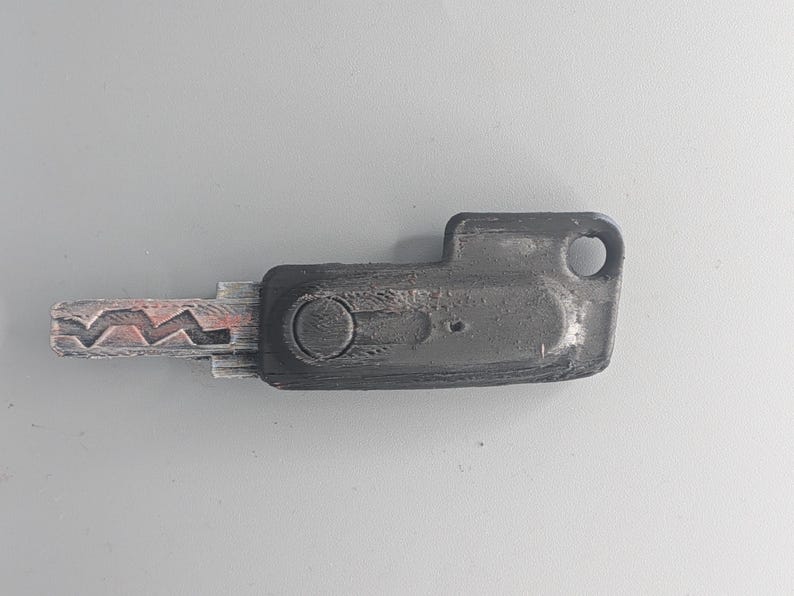 May include: A worn, dark gray key with a distinctive jagged edge and a rectangular head. The key has a circular indentation and a hole at the end, suggesting it's for a lock. The key's metal shows signs of wear.