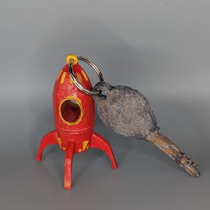May include: A red rocket-shaped keyring with yellow detailing and a silver key ring. The keyring is attached to a weathered, grey key. The rocket has three legs and a circular opening.