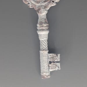 May include: An ornate, antique-style key with a detailed, decorative head and a textured, ridged handle. The key is primarily white with brown accents, suggesting an aged or weathered appearance. The key's design evokes a vintage aesthetic.