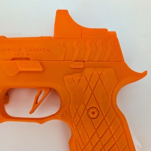 May include: Bright orange handgun with the text "ALLIGATOR SNAPPER" on the side. The gun features a trigger, grip, and sight. The grip has a diamond pattern. The gun is set against a white background.