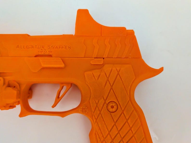 May include: Bright orange handgun with the text "ALLIGATOR SNAPPER" on the side. The gun features a trigger, grip, and sight. The grip has a diamond pattern. The gun is set against a white background.