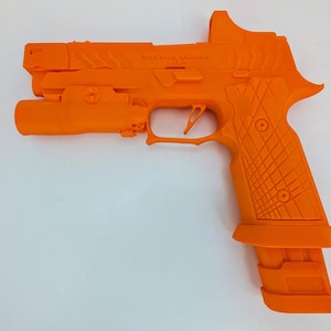 May include: Bright orange handgun with a tactical light and sight attachment. The grip features a textured pattern for enhanced handling. The text "ALLIGATOR SOLUTIONS" is visible on the slide. The magazine is inserted into the handle.
