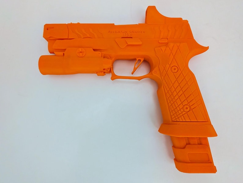 May include: Bright orange handgun with a tactical light and sight attachment. The grip features a textured pattern for enhanced handling. The text "ALLIGATOR SOLUTIONS" is visible on the slide. The magazine is inserted into the handle.