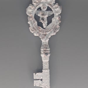 May include: A decorative key with a white, textured finish and ornate detailing. The key features a circular design with a figure in the center. The key's handle and teeth are also white, with hints of brown peeking through the finish.