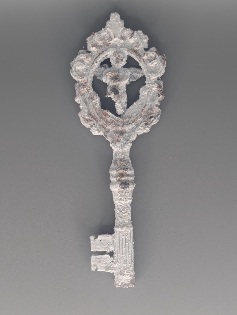 May include: A decorative key with a white, textured finish and ornate detailing. The key features a circular design with a figure in the center. The key's handle and teeth are also white, with hints of brown peeking through the finish.