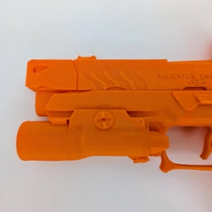 May include: Bright orange handgun with the text "ALLIGATOR SNAPPER" on the side. The firearm has a light attachment and a red dot sight. The gun is set against a white background.
