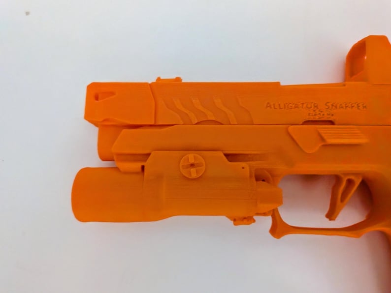 May include: Bright orange handgun with the text "ALLIGATOR SNAPPER" on the side. The firearm has a light attachment and a red dot sight. The gun is set against a white background.