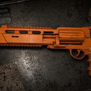 May include: A bright orange toy revolver with a long barrel, a top rail, and a textured grip featuring a wing design. The firearm replica has a weathered finish and is positioned horizontally, showcasing a large cylinder and a trigger guard.