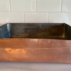 May include: A large, rectangular copper roasting pan with two brass handles. The pan is empty and has a worn, aged appearance.