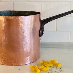 May include: A vintage copper pot with a black iron handle. The pot is sitting on a white countertop with a few pieces of yellow pasta scattered around it.