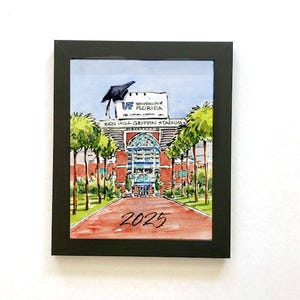 May include: A watercolor painting of the Ben Hill Griffin Stadium at the University of Florida. The stadium is framed in black and has a graduation cap on top of the sign. The year 2025 is written in black at the bottom of the painting.