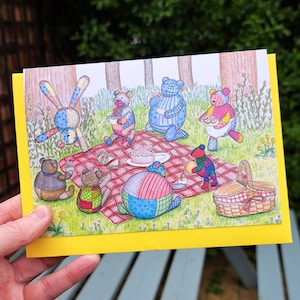 May include: A greeting card featuring a whimsical illustration of patchwork bears having a picnic in the woods. The bears are sitting around a red and white checked blanket with a cake, teapot, and other picnic items. The card has a yellow border.