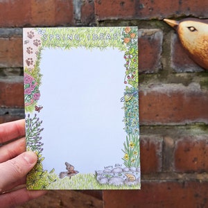 May include: A white notepad with a green, yellow, and pink floral border. The notepad has the words "SPRING IDEAS" at the top and the name "Kerry Dilks" at the bottom.