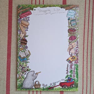 May include: A decorative shopping list notepad featuring a border of illustrated items, including baked goods, books, and art supplies. A gray mole character is shown holding a pencil. The notepad has the words "Shopping List" at the top.